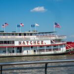 El Barco de Vapor Natchez con rueda de paletas navega en el río, ondeando banderas estadounidenses y estatales bajo un cielo azul