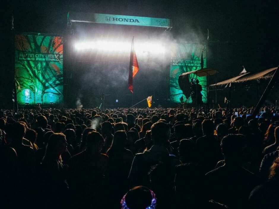 Multitud disfrutando de un concierto nocturno en el Austin City Limits Music Festival, con iluminación dramática y banderas ondeando, reflejando la vibrante atmósfera del evento