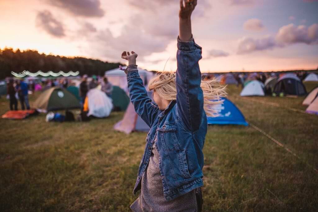 Mujer joven con chaqueta de mezclilla celebrando al atardecer en un festival de música, con carpas coloridas y público al fondo en un ambiente relajado y festivo