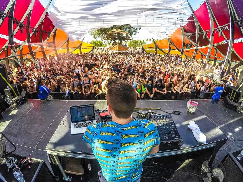 Vista desde detrás de un DJ mirando hacia una multitud entusiasta bajo una estructura colorida en un día soleado, capturando la energía vibrante de un festival de música