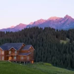 Una cabaña de madera en primer plano, con montañas al atardecer en el fondo y un bosque de pinos en un paisaje tranquilo