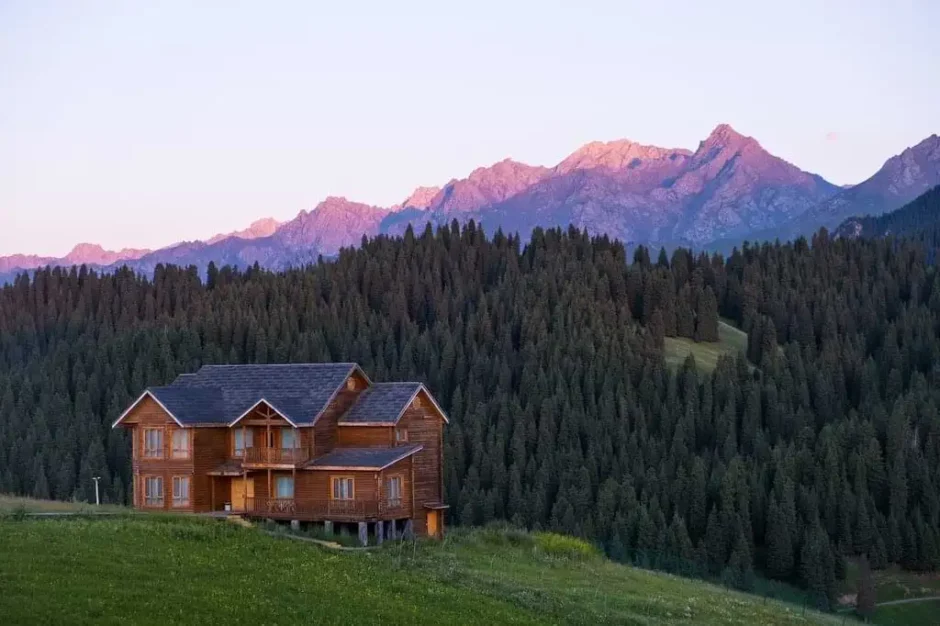 Una cabaña de madera en primer plano, con montañas al atardecer en el fondo y un bosque de pinos en un paisaje tranquilo