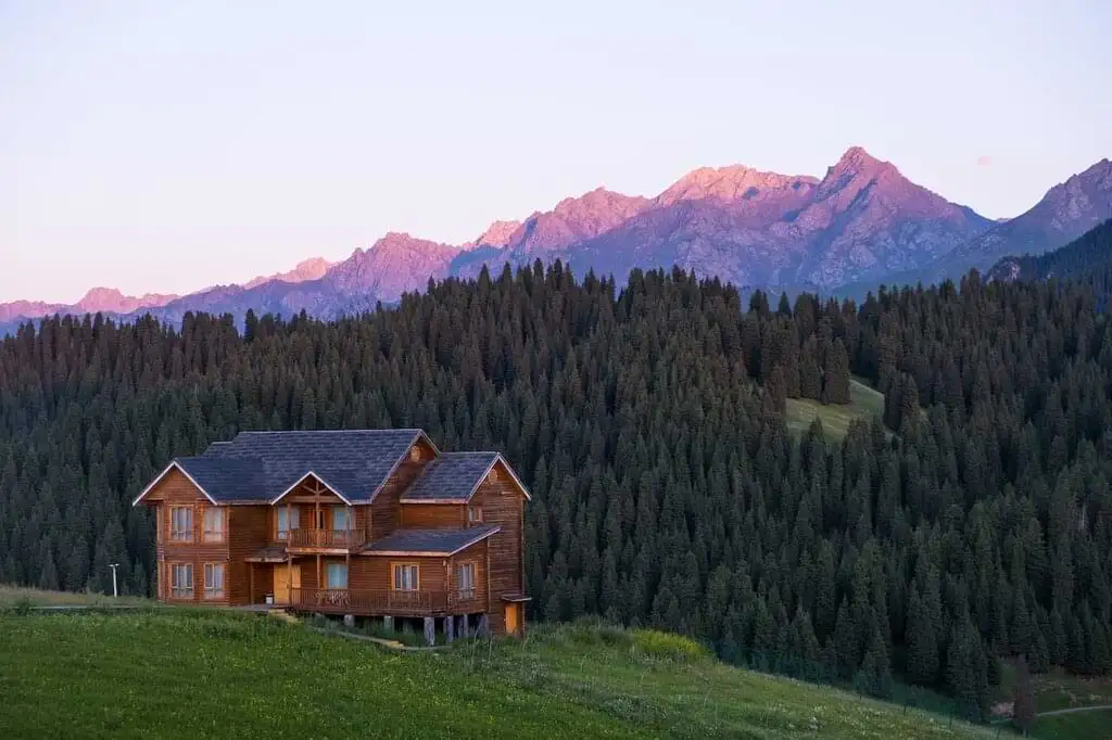 Una cabaña de madera en primer plano, con montañas al atardecer en el fondo y un bosque de pinos en un paisaje tranquilo