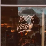 Neón con el texto '100% VEGAN' destaca en la ventana de un restaurante, invitando a los transeúntes a explorar opciones alimenticias compasivas y sostenibles