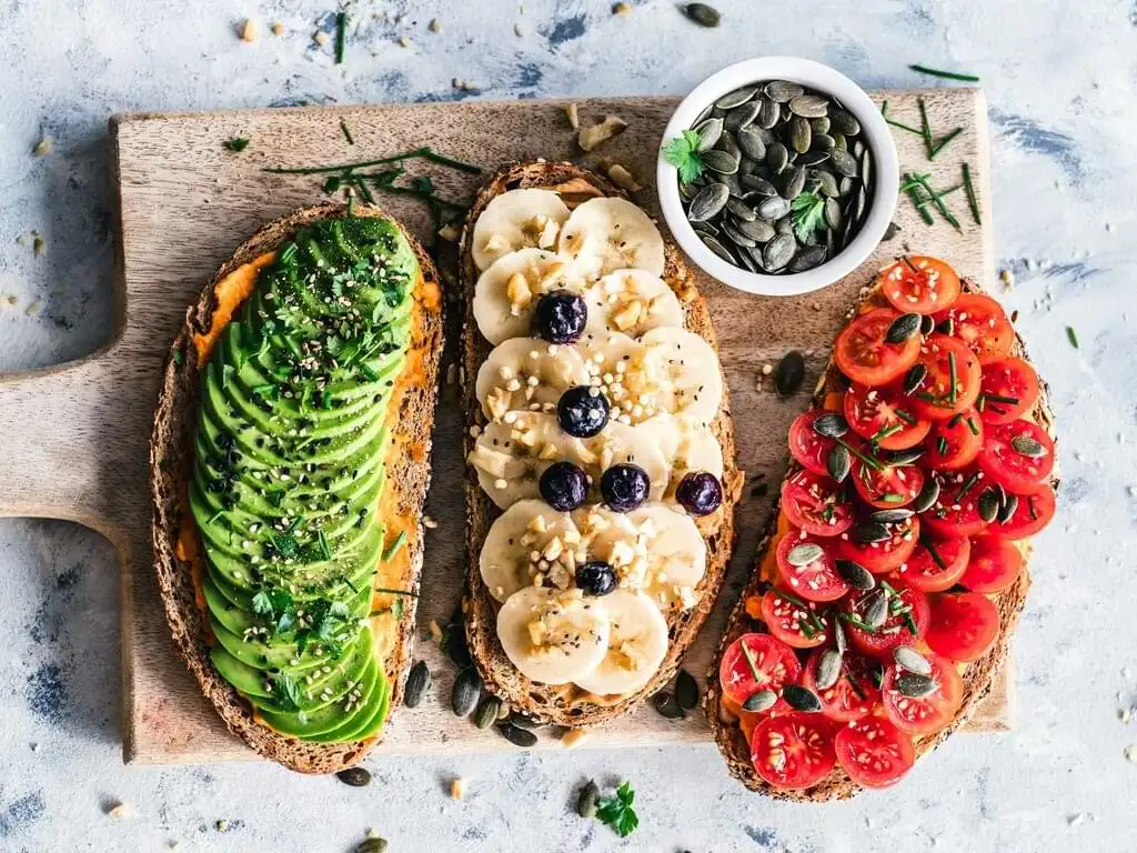 Tres tostadas veganas con aguacate, plátano y bayas, y tomate sobre una tabla de madera, acompañadas de un bol de semillas de calabaza