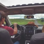 Conductor en primer plano manejando por una carretera despejada con cielo nublado, acompañado de sombreros en el asiento del copiloto, evocando un viaje de exploración y aventura