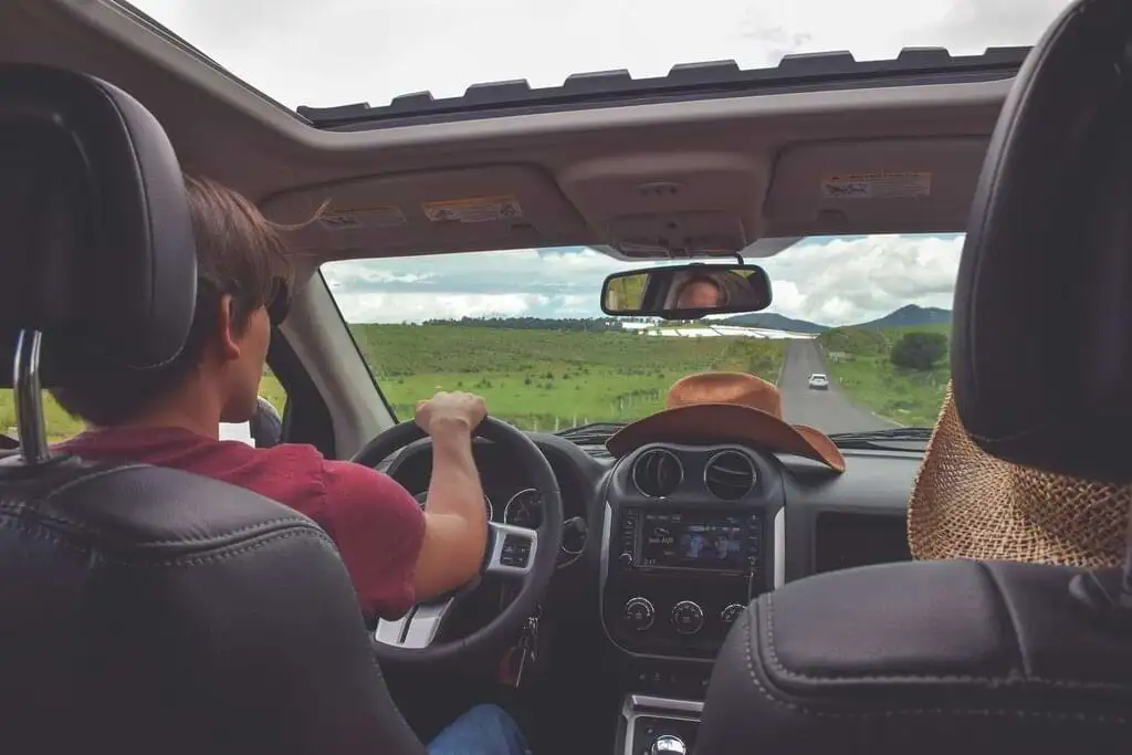 Conductor en primer plano manejando por una carretera despejada con cielo nublado, acompañado de sombreros en el asiento del copiloto, evocando un viaje de exploración y aventura