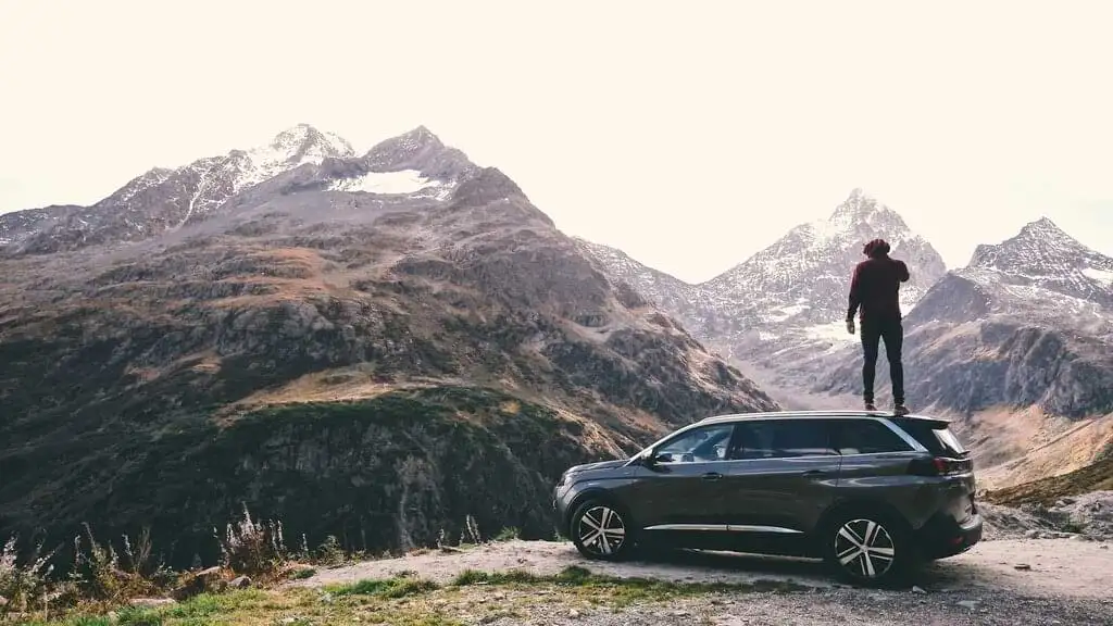 Persona de pie sobre el techo de un coche, contemplando montañas nevadas, capturando la esencia de la aventura y la inmensidad de la naturaleza