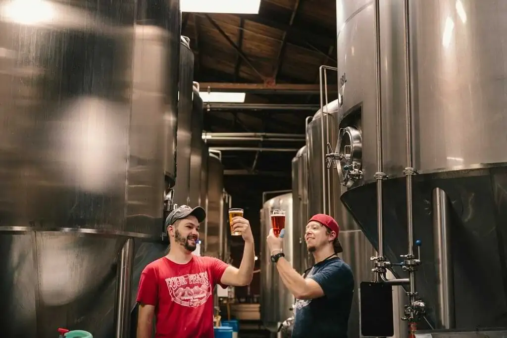 Dos cerveceros con gorras brindando alegremente, uno examinando una pinta de cerveza, con tanques de acero inoxidable al fondo