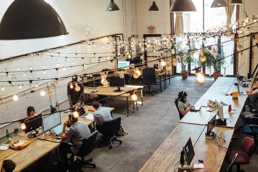 Profesionales concentrados trabajando en un amplio y moderno espacio de coworking adornado con luces colgantes y plantas, fomentando un ambiente productivo