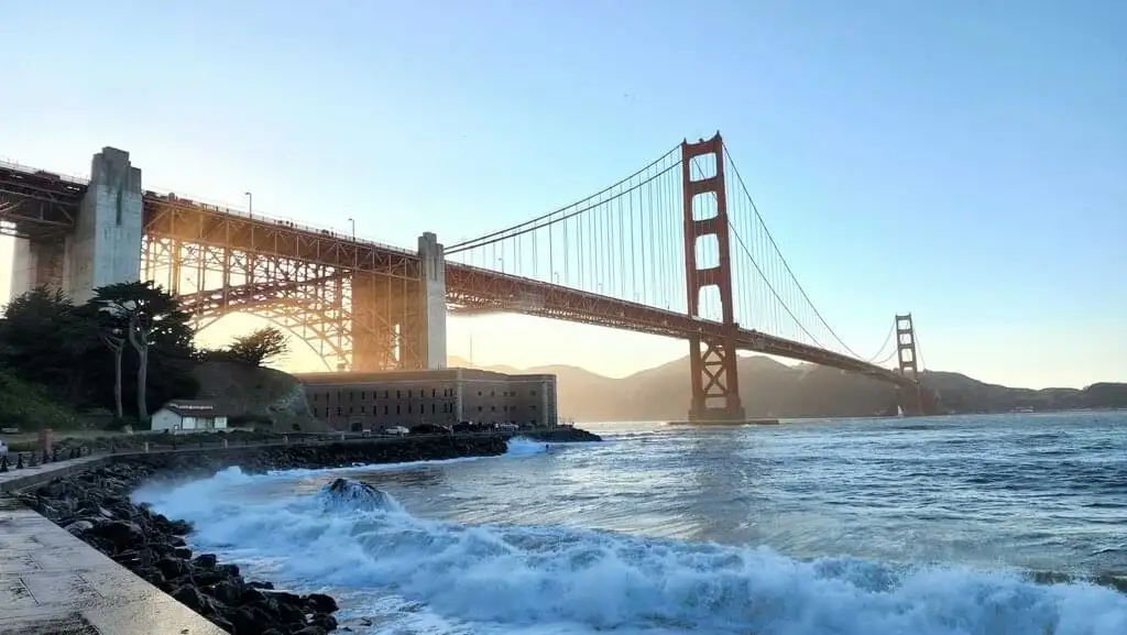 Vista del Golden Gate Bridge en San Francisco durante el atardecer, con olas chocando contra la orilla y un cielo despejado