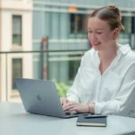 Una joven sonriente trabajando en dos laptops al aire libre en un espacio urbano moderno, representando el estilo de vida de un trabajador remoto o nómada digital