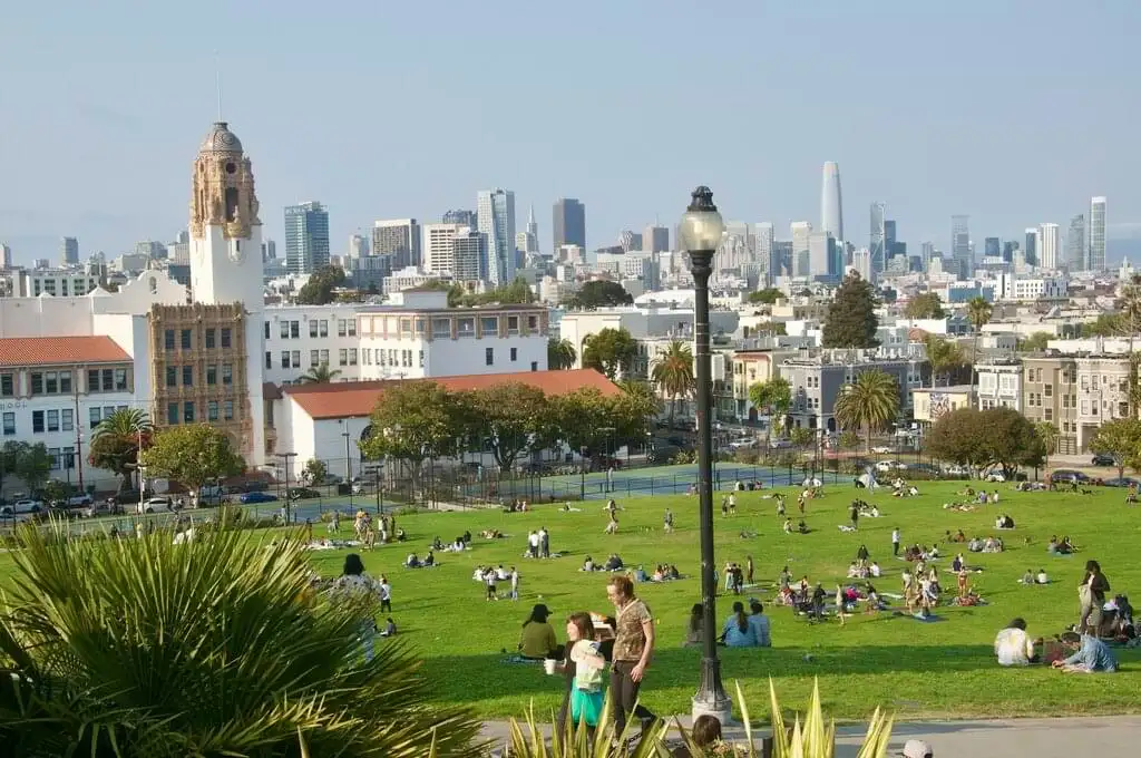 Gente relajándose en un parque soleado con la silueta urbana de San Francisco al fondo, destacando arquitectura histórica y moderna