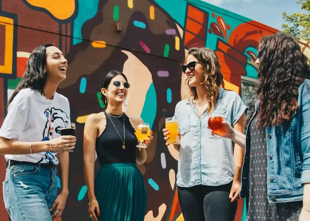 Grupo de amigos sonrientes disfrutando de cervezas al aire libre, con un mural colorido de arte urbano de fondo