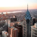 Vista aérea cautivadora del skyline de Filadelfia durante el atardecer. Edificios prominentes incluyen el ornamentado Ayuntamiento de Filadelfia con su icónica torre de reloj y rascacielos modernos, especialmente Liberty Place con su cima en forma de pirámide. La ciudad se extiende hacia el horizonte, y un río brilla a lo lejos