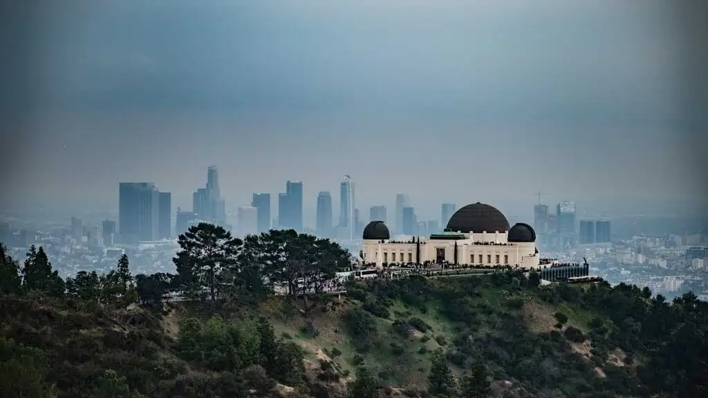 Vista matutina del Observatorio Griffith en Los Ángeles, ubicado en una colina verde, con el centro de la ciudad en el fondo. El cielo está nublado y la ciudad se ve envuelta en una ligera neblina, creando una atmósfera tranquila y serena