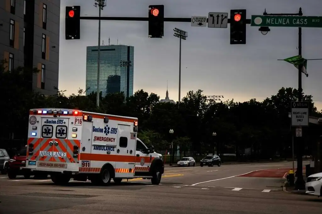 Una ambulancia de emergencia de Boston EMS avanzando por una intersección de la ciudad con las luces de emergencia encendidas. Al fondo se observa el paisaje urbano con edificios altos, árboles y señales de tránsito bajo un cielo nublado al atardecer