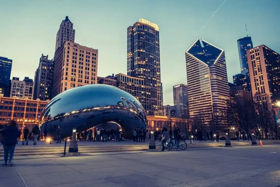 Fotografía de un anochecer en Chicago que muestra el icónico Cloud Gate, conocido como el Bean, reflejando los edificios iluminados del centro de la ciudad. En primer plano, personas pasean y disfrutan del ambiente urbano, mientras los rascacielos se alzan en el fondo bajo un cielo azul oscuro