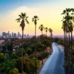 Vista panorámica al atardecer de Los Ángeles, mostrando una carretera flanqueada por altas palmeras, con el centro de la ciudad en el horizonte bajo un cielo despejado y un pequeño cuarto creciente de luna visible