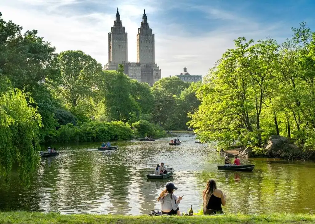 Personas disfrutando de un día soleado en Central Park, Nueva York. Varias personas están en botes en el lago rodeado de exuberante vegetación y árboles. Dos mujeres sentadas en la orilla del lago conversan y observan el paisaje, con edificios icónicos al fondo