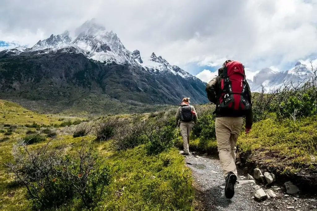 Dos excursionistas con mochilas grandes caminando por un sendero de montaña rodeado de vegetación. Al fondo se pueden ver montañas con picos nevados y el cielo parcialmente nublado, creando un entorno natural impresionante y aventurero