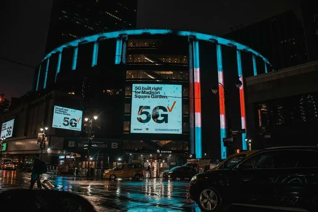 Vista nocturna de la fachada del Madison Square Garden iluminada con luces azules y rojas. Los anuncios de tecnología 5G son visibles en las pantallas exteriores del edificio. La escena incluye tráfico y peatones bajo la luz de las farolas en una noche lluviosa en la ciudad.