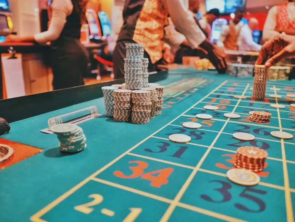 Mesa de ruleta en un casino de Las Vegas con fichas apiladas y dispersas sobre los números. Varios jugadores y crupieres en el fondo. El ambiente muestra la actividad y emoción del juego de azar en un casino iluminado y animado.