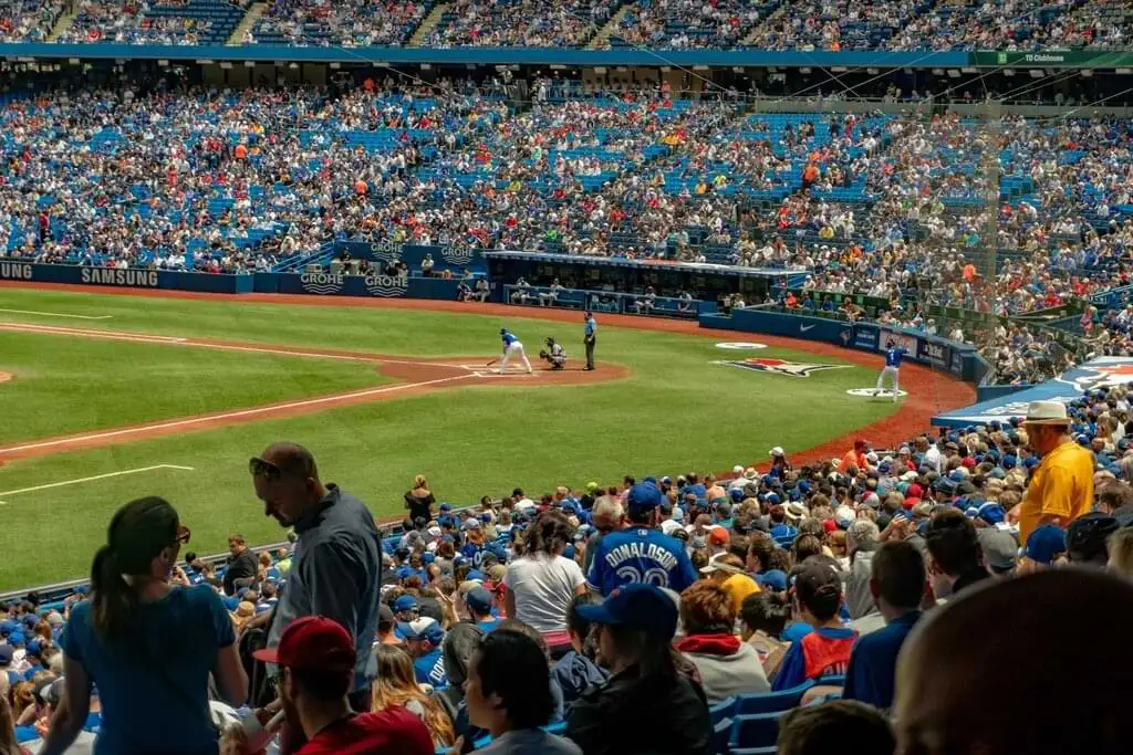 Vista desde las gradas de un estadio de béisbol durante un partido. Los aficionados llenan las gradas mientras un bateador se prepara en el plato. Se observan las áreas de los dugouts y el campo de juego, con un ambiente animado y lleno de emoción deportiva.