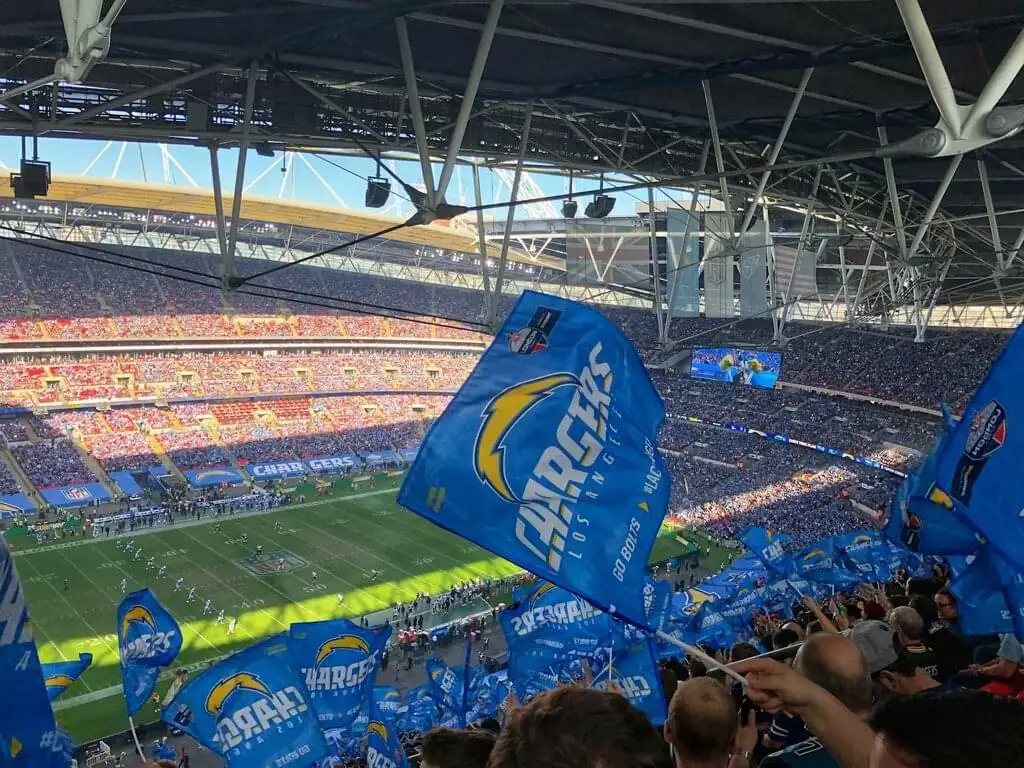 Vista desde las gradas de un estadio de fútbol americano lleno de espectadores. Los aficionados ondean banderas del equipo Los Angeles Chargers. Se puede ver el campo de juego y una pantalla gigante mostrando el partido. El estadio está lleno de energía y emoción.