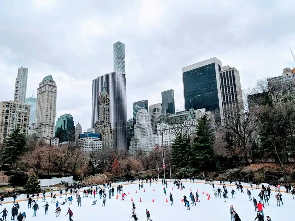 Personas disfrutando de una pista de patinaje sobre hielo en Central Park, Nueva York, rodeadas de rascacielos que forman parte del icónico skyline de la ciudad. La escena muestra un día nublado de invierno con árboles sin hojas alrededor de la pista