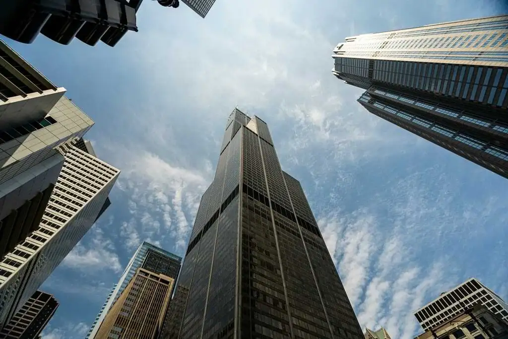 Fotografía tomada desde el suelo mirando hacia arriba, mostrando varios rascacielos en Chicago que se elevan hacia un cielo parcialmente nublado. La imagen captura la imponencia de la arquitectura moderna y la grandeza de los edificios que definen el horizonte de la ciudad