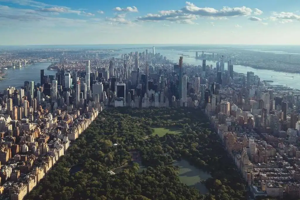 Vista aérea de Central Park en Nueva York con el impresionante skyline de Manhattan en el fondo. Se pueden apreciar los altos rascacielos rodeando el parque y el río Hudson a la izquierda. El cielo despejado con algunas nubes añade un toque sereno a la escena urbana