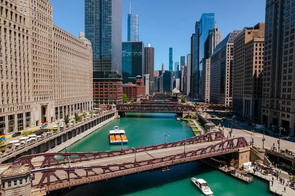 Imagen del Río Chicago en un día soleado, con varios puentes que cruzan el río y altos edificios a ambos lados. Una pequeña embarcación y un taxi acuático navegan por las aguas color turquesa, mientras la arquitectura urbana se destaca contra el cielo azul