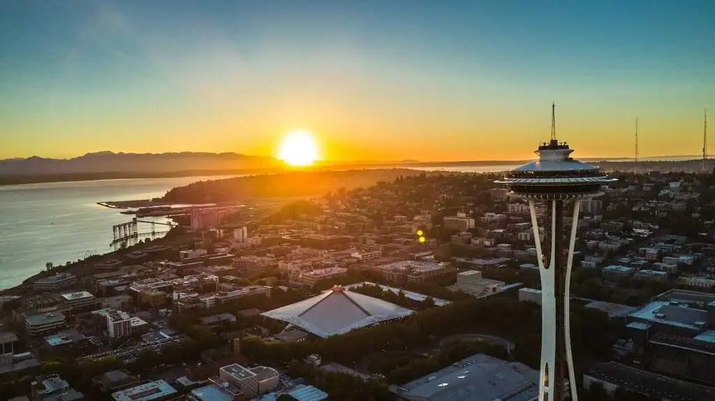 Vista aérea del atardecer sobre Seattle, destacando la Space Needle en primer plano y el sol descendiendo detrás de las colinas, iluminando el horizonte y el agua del puerto con tonos dorados.