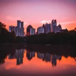 Una hermosa vista del atardecer sobre el skyline de Atlanta, con el cielo teñido de tonos rosados y anaranjados reflejándose en el agua tranquila de un lago, enmarcado por árboles oscuros a ambos lados.