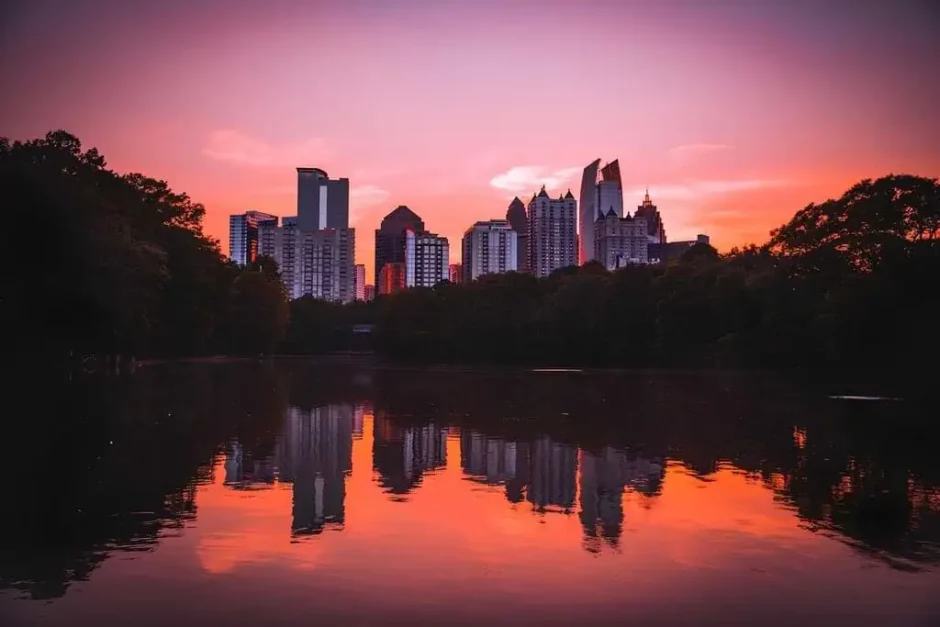 Una hermosa vista del atardecer sobre el skyline de Atlanta, con el cielo teñido de tonos rosados y anaranjados reflejándose en el agua tranquila de un lago, enmarcado por árboles oscuros a ambos lados.