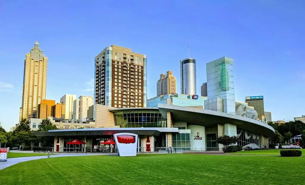 Vista del Centro de Coca-Cola en Atlanta con modernos edificios de gran altura al fondo bajo un cielo despejado. El edificio principal del centro destaca con su diseño contemporáneo y su famoso logotipo rojo, rodeado de un verde césped bien cuidado.