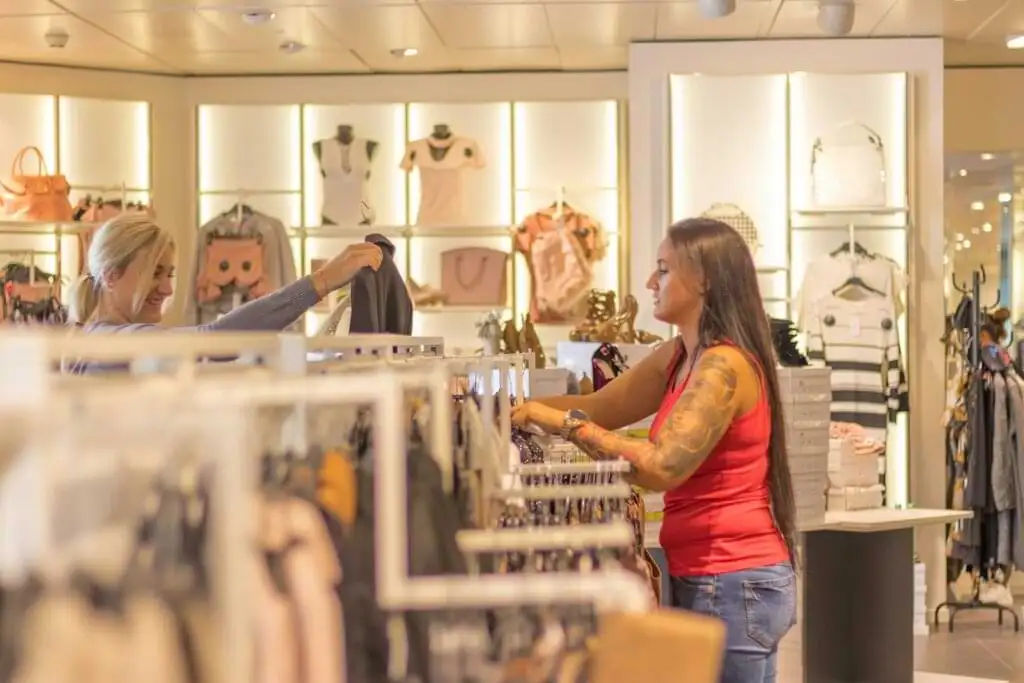 Dos mujeres comprando en una tienda de moda, mirando ropa y accesorios. La tienda está bien iluminada, con estantes y exhibiciones de ropa y bolsos. Una de las mujeres muestra una prenda a la otra mientras sonríen y disfrutan de la experiencia de compra.