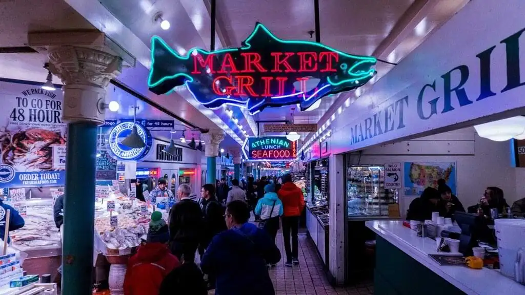 "Vista del interior del Pike Place Market en Seattle, mostrando el Market Grill y otros puestos de mariscos con letreros de neón, y varias personas recorriendo el mercado mientras compran productos frescos."