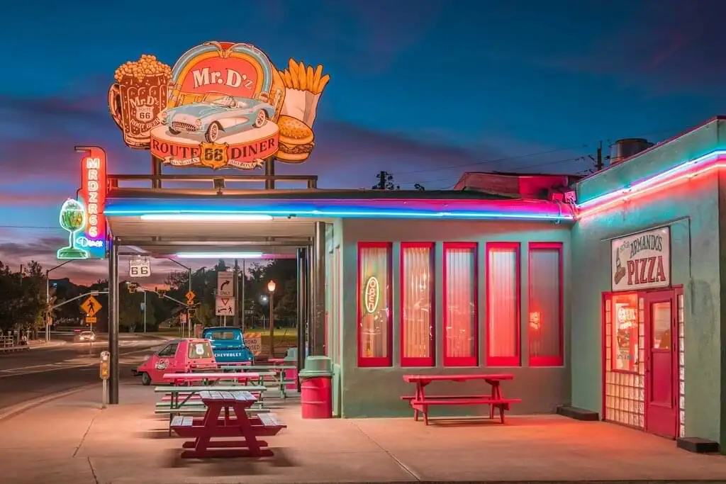 Vista nocturna del restaurante Mr. D'z Route 66 Diner, un icónico establecimiento retro con neones en Kingman, Arizona. El diner presenta un letrero luminoso con un coche clásico y una decoración de los años 50. En la entrada hay mesas de picnic y un auto antiguo estacionado, todo bajo un cielo crepuscular.