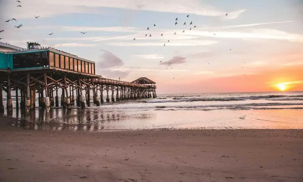 Vista del muelle de Cocoa Beach en Florida durante el atardecer. El sol se pone en el horizonte mientras las aves vuelan sobre el mar y las olas rompen suavemente en la playa. El muelle se extiende hacia el agua con una estructura de madera.