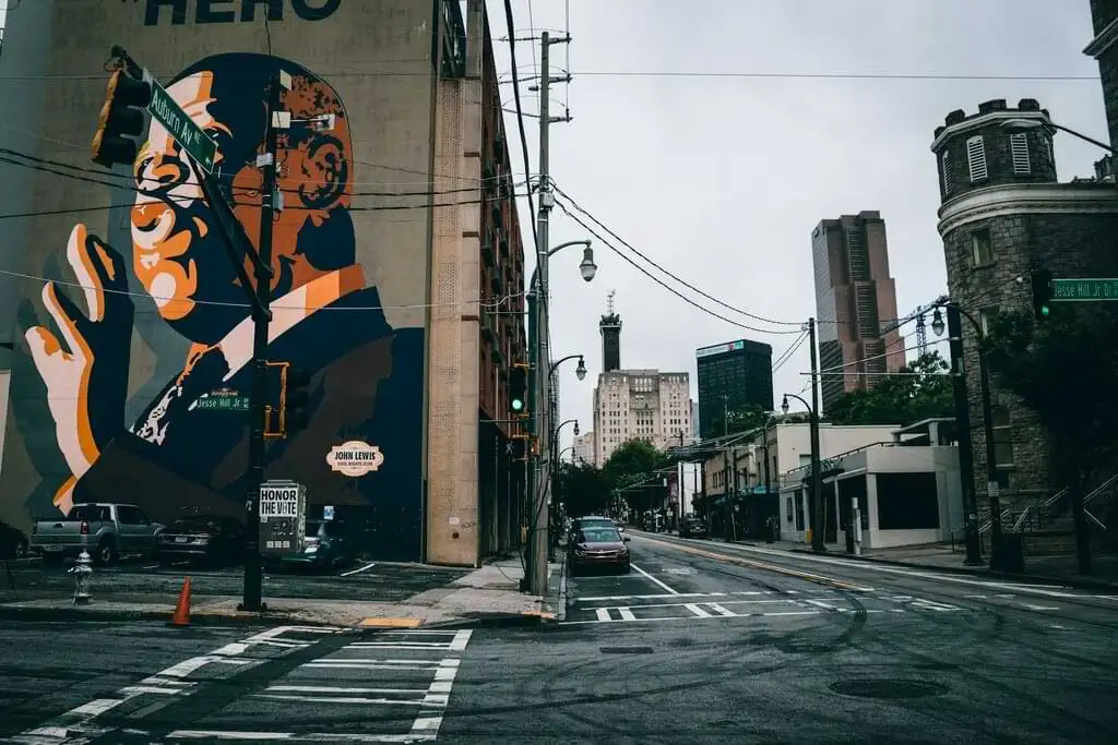 Mural de John Lewis en una calle de Atlanta, con un diseño artístico que resalta su perfil y la palabra "HERO". La calle está tranquila con pocos coches, y edificios históricos y modernos se mezclan en el paisaje urbano bajo un cielo nublado.