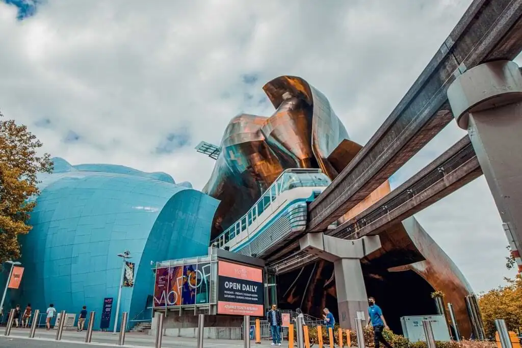 "Vista del Museo de la Cultura Pop (MoPOP) en Seattle, destacando su arquitectura moderna y colorida, con el monorraíl pasando por el edificio, bajo un cielo parcialmente nublado."