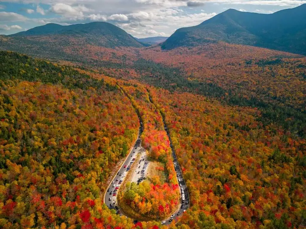 Vista aérea del pintoresco paisaje otoñal en las Montañas Blancas de New Hampshire. La carretera serpentea a través del bosque densamente cubierto de árboles que muestran una vibrante gama de colores otoñales, desde rojos y naranjas hasta amarillos. Las montañas se elevan en el horizonte bajo un cielo parcialmente nublado.