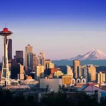 Vista panorámica del skyline de Seattle al atardecer, destacando la Space Needle y el Monte Rainier nevado en el fondo, con un cielo azul claro y edificios iluminados por la luz del sol