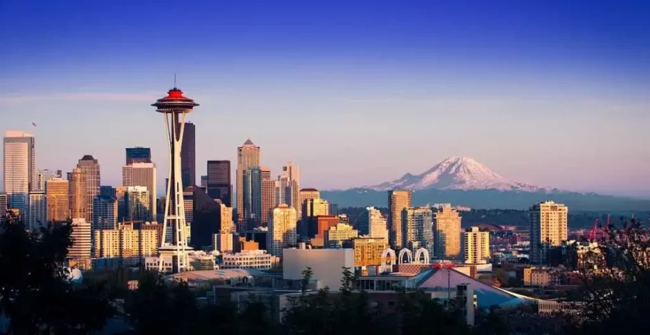 Vista panorámica del skyline de Seattle al atardecer, destacando la Space Needle y el Monte Rainier nevado en el fondo, con un cielo azul claro y edificios iluminados por la luz del sol