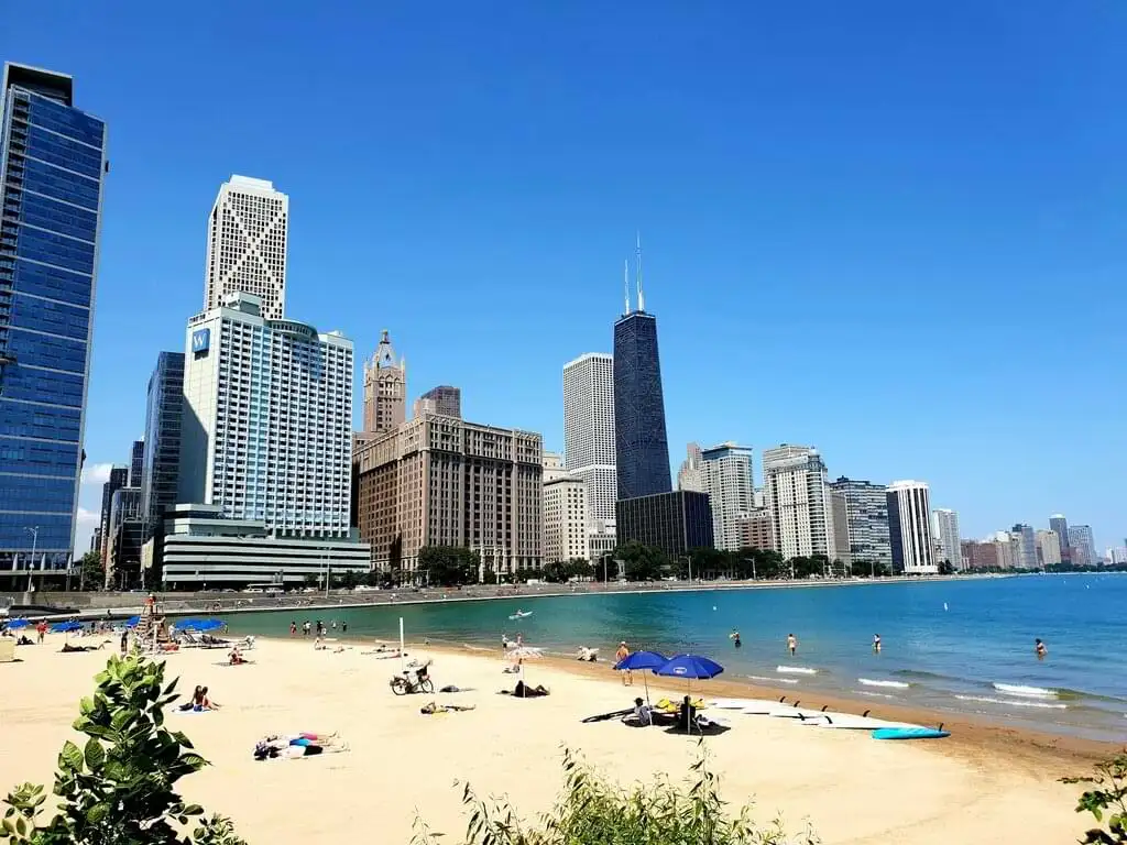 Playa urbana de Chicago junto al lago Michigan, con personas disfrutando del sol y el agua, y el impresionante skyline de la ciudad de fondo, incluyendo rascacielos icónicos como el John Hancock Center