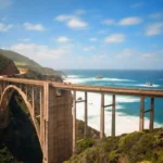 Vista panorámica del icónico Puente Bixby Creek en la costa de Big Sur, California, con el océano Pacífico de fondo. El puente de arco de hormigón se extiende majestuosamente sobre un profundo cañón, rodeado de vegetación y acantilados escarpados, en un día soleado con cielo azul y nubes dispersas.