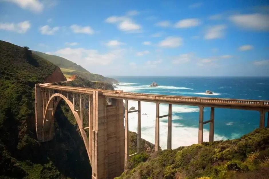 Vista panorámica del icónico Puente Bixby Creek en la costa de Big Sur, California, con el océano Pacífico de fondo. El puente de arco de hormigón se extiende majestuosamente sobre un profundo cañón, rodeado de vegetación y acantilados escarpados, en un día soleado con cielo azul y nubes dispersas.