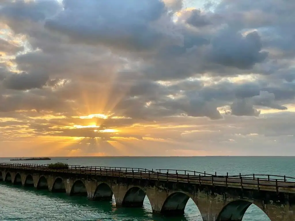 Impresionante amanecer sobre el Puente de las Siete Millas en los Cayos de Florida. Los rayos del sol emergen entre las nubes, iluminando el cielo con tonos dorados y anaranjados mientras se reflejan en las aguas tranquilas del océano. El puente antiguo, con su estructura de arcos, se extiende sobre el mar, creando una escena serena y majestuosa.